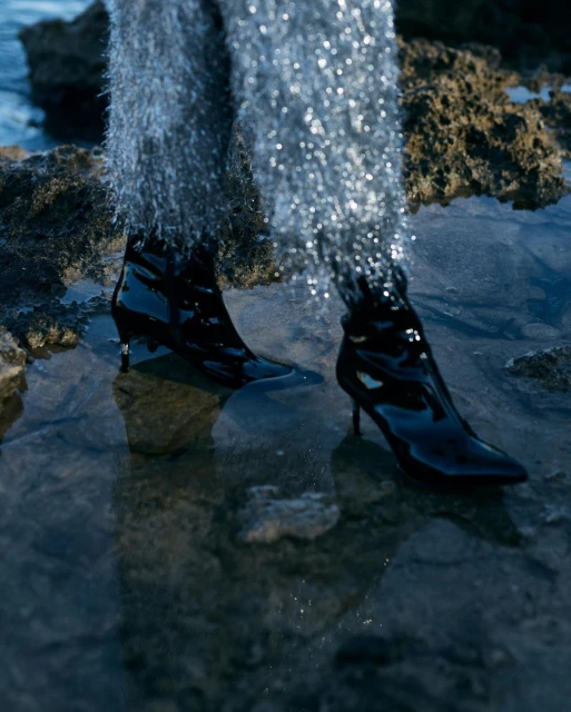 Person in shiny pants and high-heeled patent boots standing in shallow water surrounded by rocks and the sea, fashion editorial.