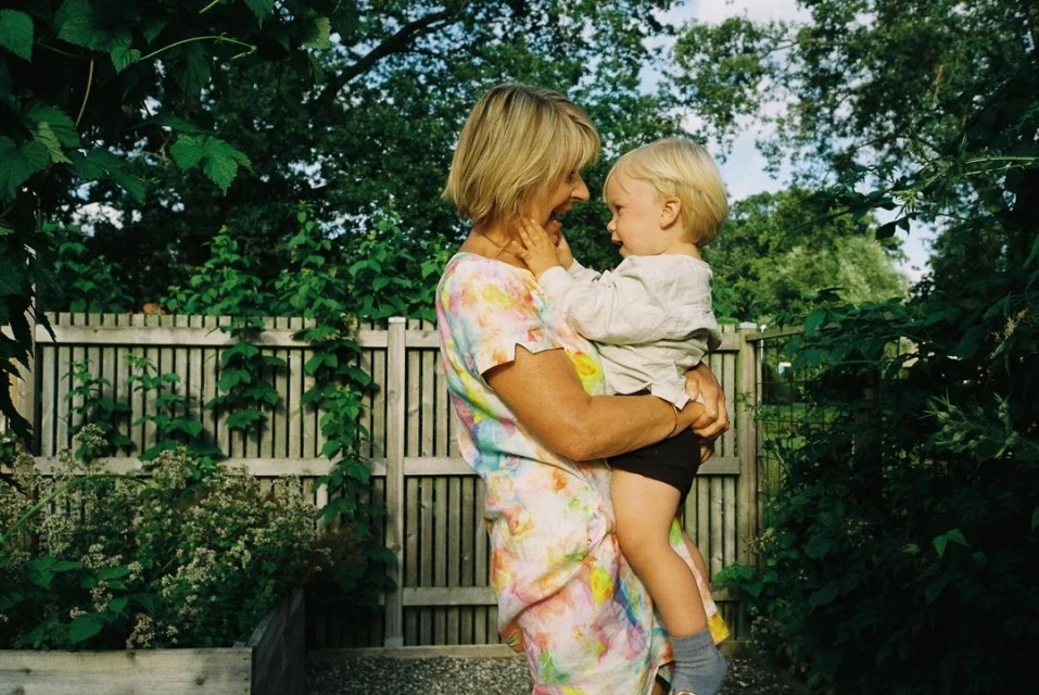 Outdoor scene featuring an adult female holding a toddler, both smiling. The background is lush with greenery, trees, and a wooden fence.