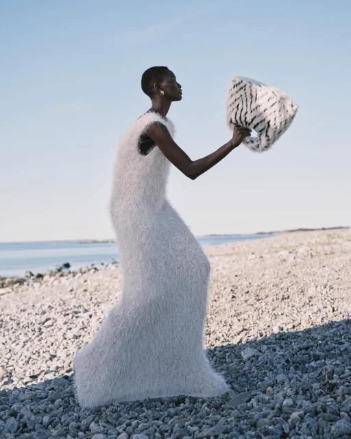 A slender Black woman in a white textured dress poses on a rocky beach, holding a patterned bag against a clear sky and ocean backdrop.