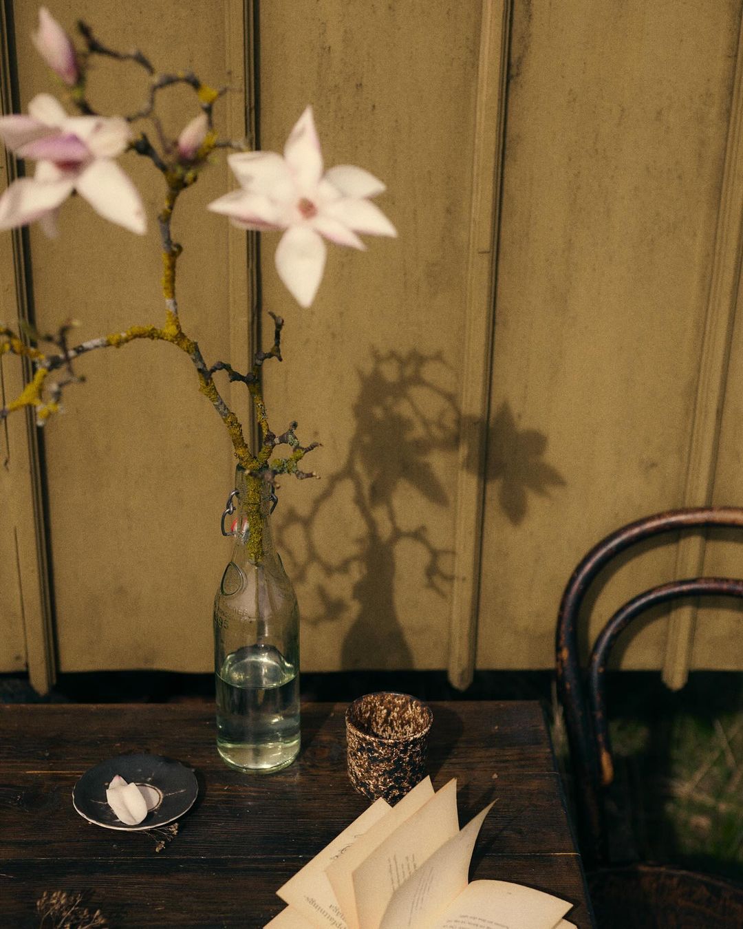 Close-up of a wooden table featuring a glass bottle vase with magnolia branches, an open book, and decorative candle holder.