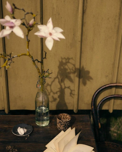 Close-up of a wooden table featuring a glass bottle vase with magnolia branches, an open book, and decorative candle holder.