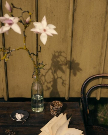 Close-up of a wooden table featuring a glass bottle vase with magnolia branches, an open book, and decorative candle holder.