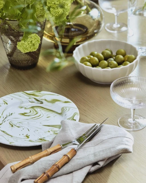 Table setting with green olives, textured plate, linen napkin, glasses and decorative vase, creating an elegant dining scene.