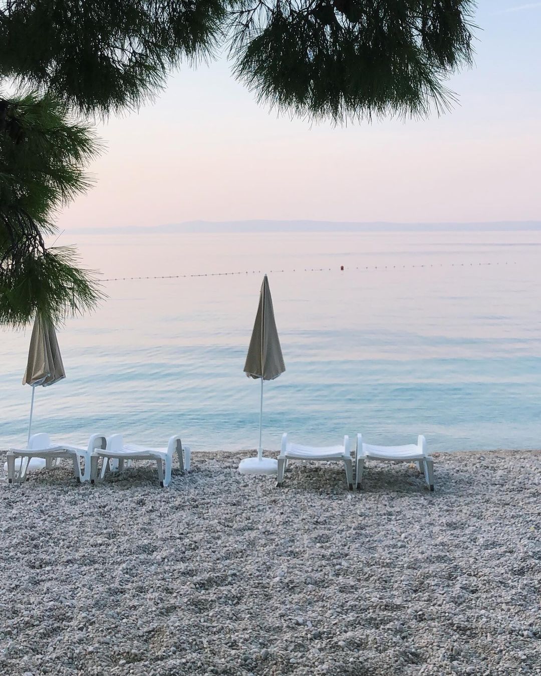 Pebble beach scene with lounge chairs and umbrellas facing the ocean at sunset with peaceful, relaxing atmosphere; vacation vibes.