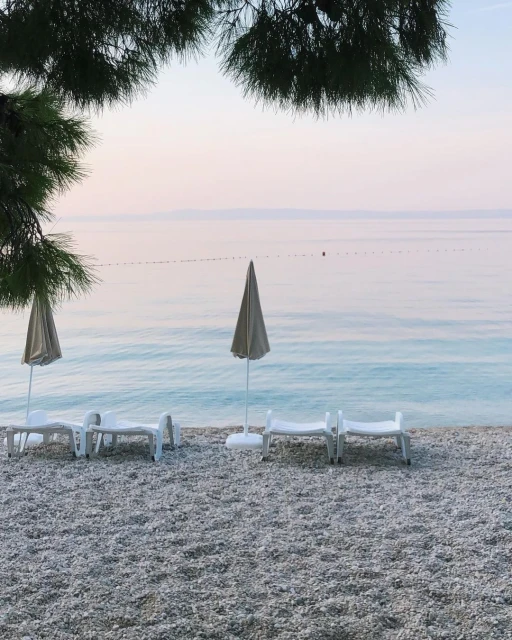 Pebble beach scene with lounge chairs and umbrellas facing the ocean at sunset with peaceful, relaxing atmosphere; vacation vibes.