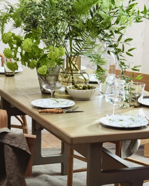 A styled dining table features floral arrangements in vases, set with plates, cutlery, glassware, and a bowl of green olives.