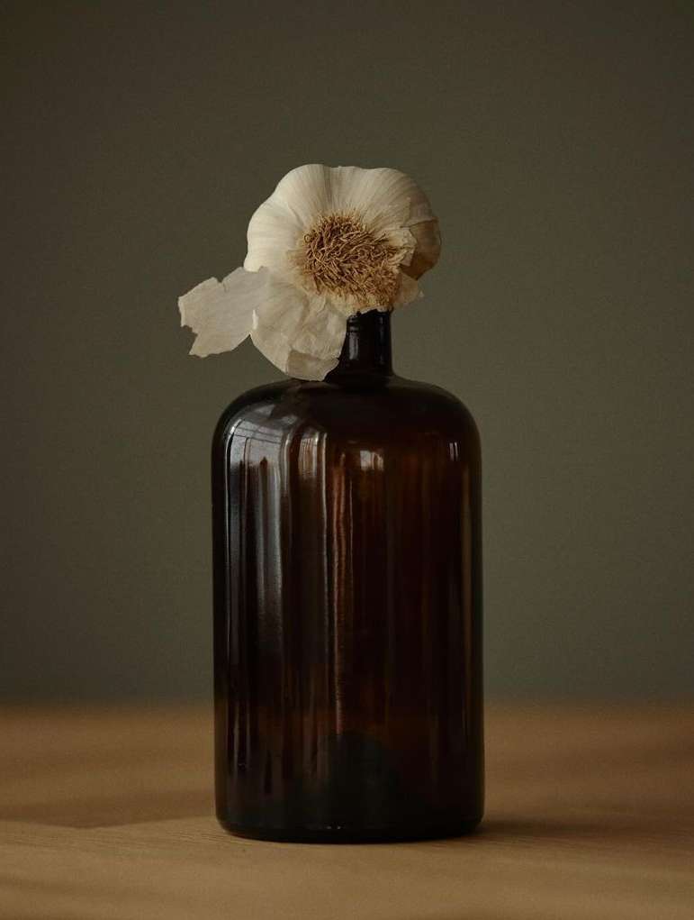 Still life shot of a garlic bulb atop a vintage brown glass bottle set on a wooden table against a neutral grey background.