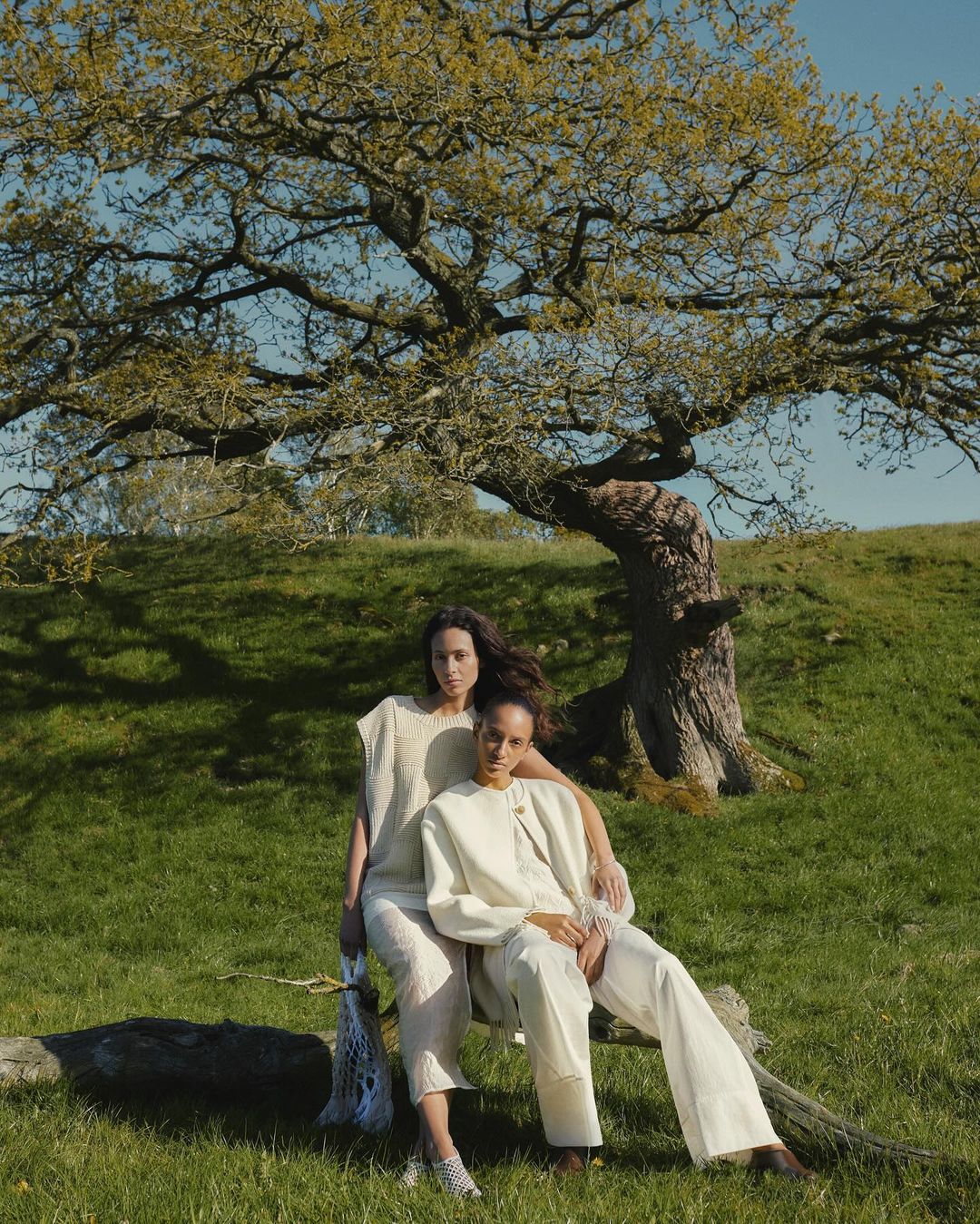 Two women pose in nature wearing matching neutral-toned outfits, seated on a log with a large leafy tree in the background.