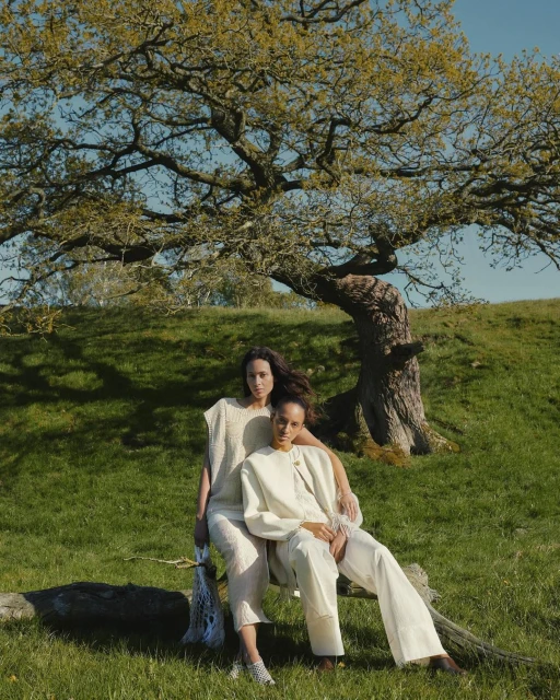Two women pose in nature wearing matching neutral-toned outfits, seated on a log with a large leafy tree in the background.