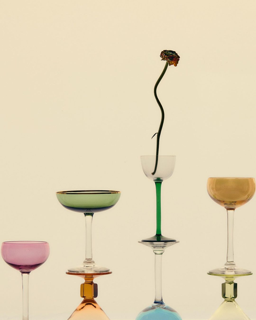 Still life with colorful vintage coupe glasses stacked in a staggered row, one holding a single withered flower, cream background, studio lighting.