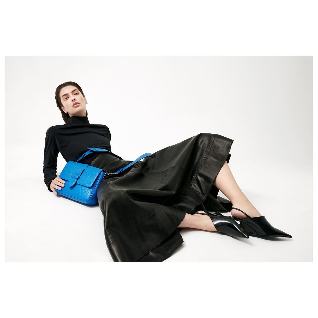 Woman in black turtleneck and skirt lying down holding a bright blue purse in a studio environment. Fashion and style concept.