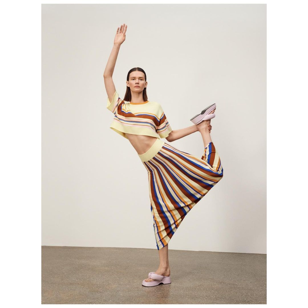An adult woman poses in a yellow and brown striped knitwear skirt and crop top with platform sandals against a neutral backdrop.