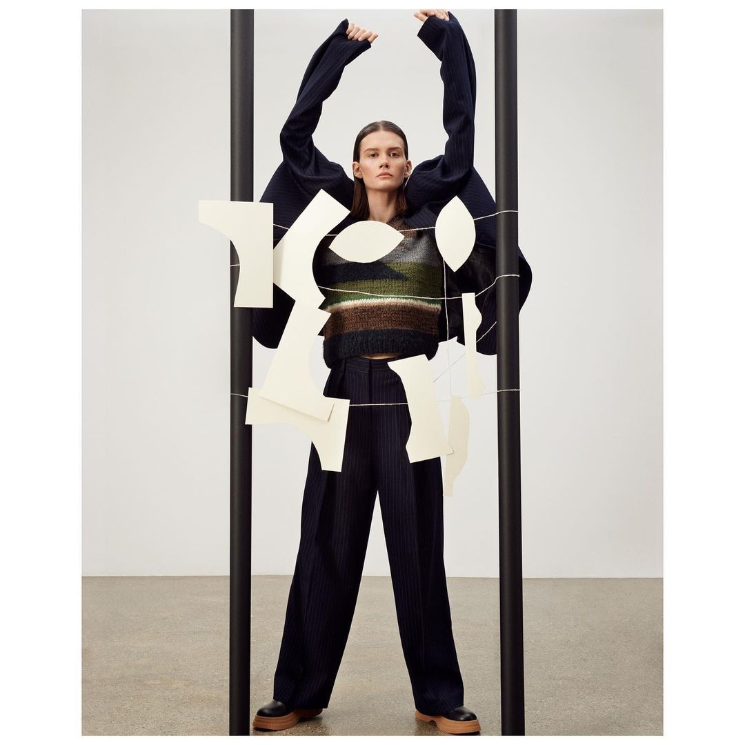 A woman in a patterned sweater, dark wide-leg pants, and a boxy jacket poses with geometric paper cutouts in a fashion studio setting.