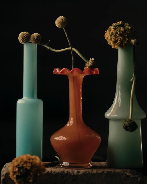 Three pastel-colored glass vases of different shapes hold dried, round flowers against a dark background in a still life composition.