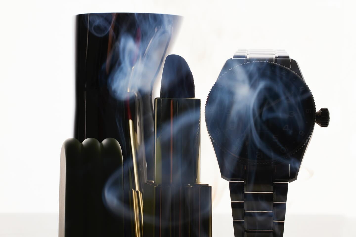 Studio shot of beauty items: lipstick, face stone, and watch. Vaporous smoke swirls in the shot. Minimalist design on white.