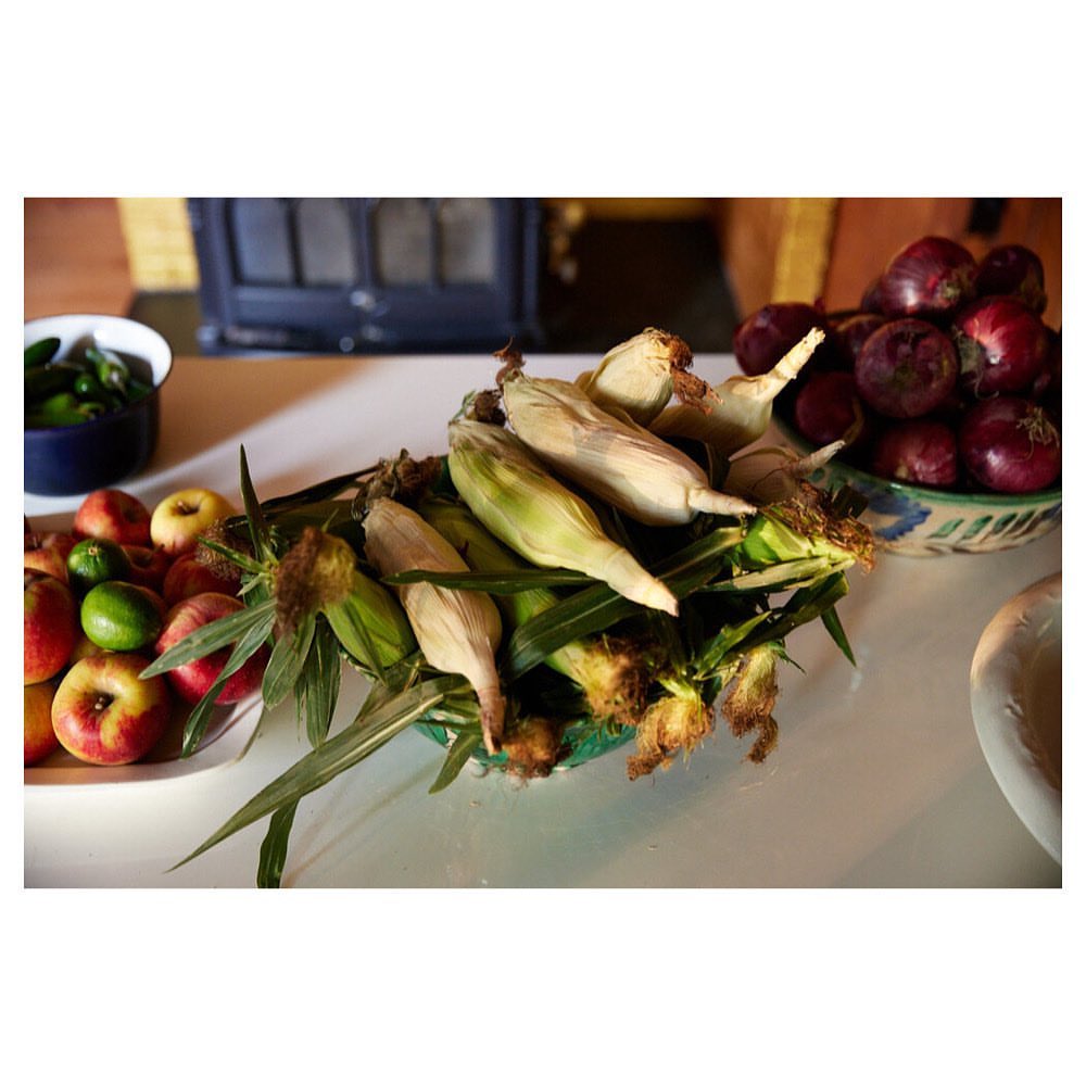 Overhead shot of fresh corn on the cob in a ceramic bowl on a white table, with a variety of fruits and vegetables displayed nearby.