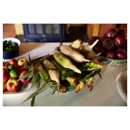 Overhead shot of fresh corn on the cob in a ceramic bowl on a white table, with a variety of fruits and vegetables displayed nearby.