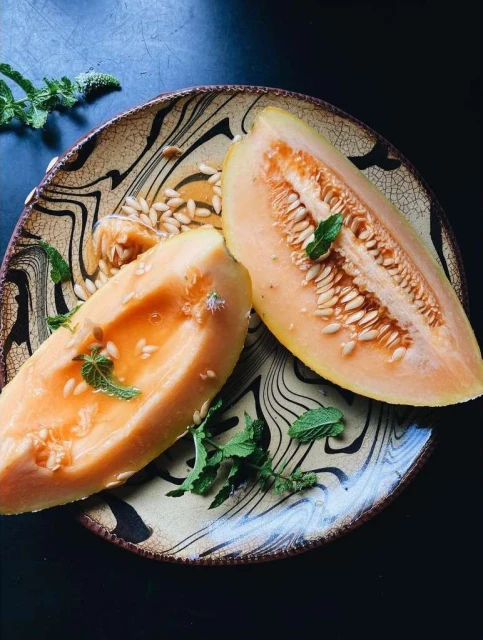 Overhead shot of cantaloupe slices on an earth-toned plate, garnished with mint leaves, set against a dark background.