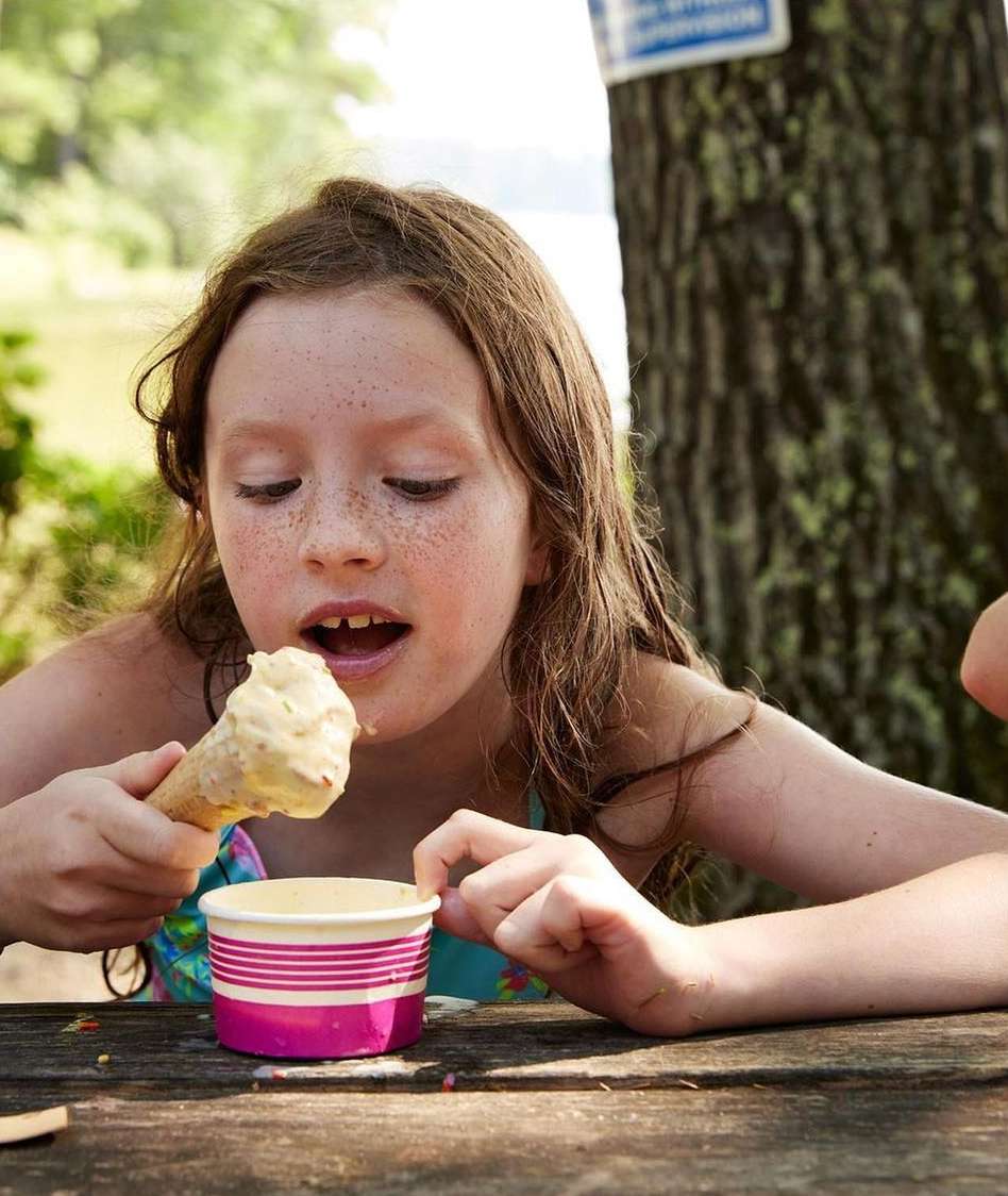 A young girl with freckles eats ice cream from a cone and a cup at an outdoor picnic table on a sunny day near trees and a lake.