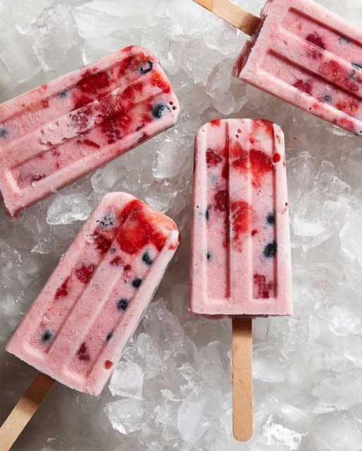 Four berry ice pops on sticks, arranged on a bed of crushed ice, featuring a pink hue and a refreshing summer treat aesthetic.