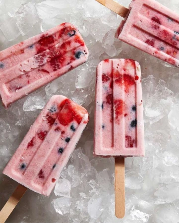 Four berry ice pops on sticks, arranged on a bed of crushed ice, featuring a pink hue and a refreshing summer treat aesthetic.