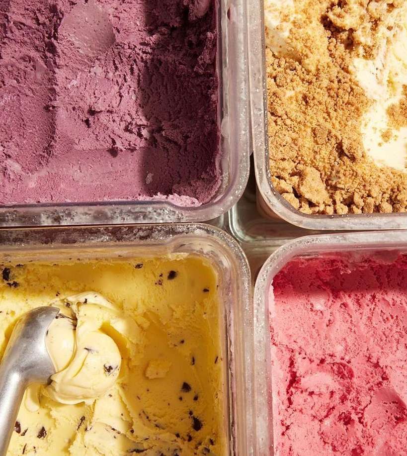 Overhead shot of colorful ice cream in clear containers, featuring purple, beige, yellow, and pink flavors, highlighted by natural light.