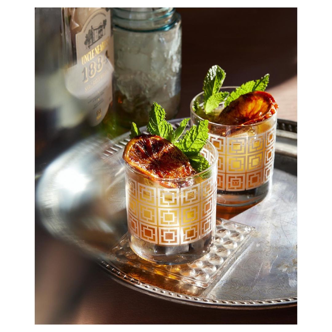 Two cocktails garnished with mint sprigs and caramelized orange slices sit on a silver tray next to a liquor bottle and glass jar.