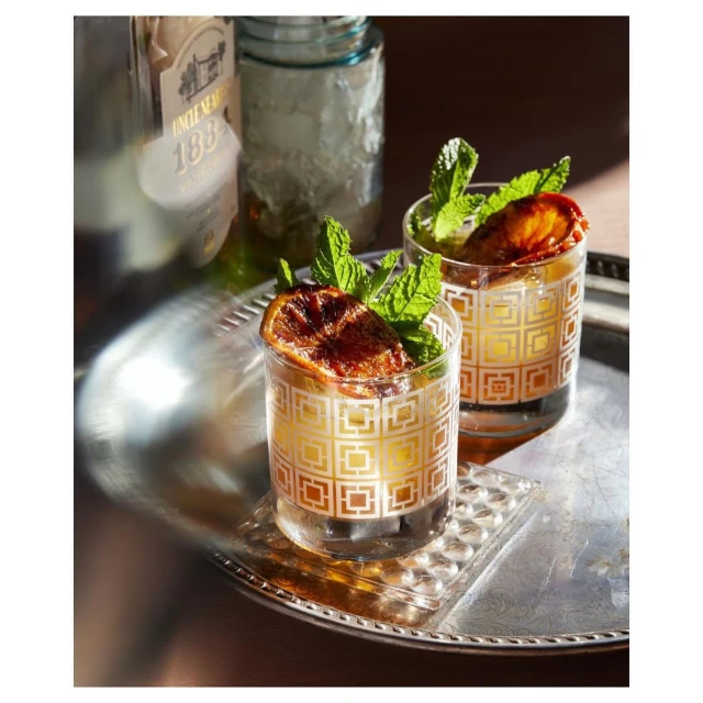 Two cocktails garnished with mint sprigs and caramelized orange slices sit on a silver tray next to a liquor bottle and glass jar.