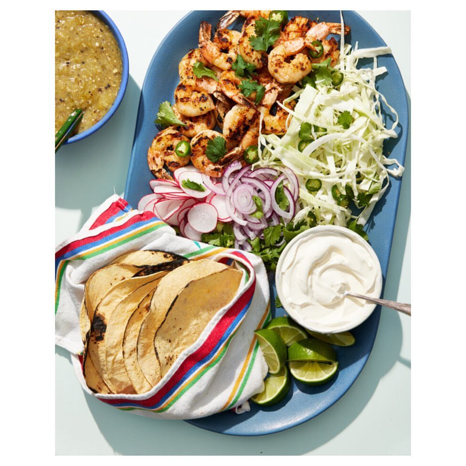 Overhead shot of grilled shrimp on a blue platter, accompanied by cabbage, lime, radishes, red onions, tortillas, sour cream, and salsa.