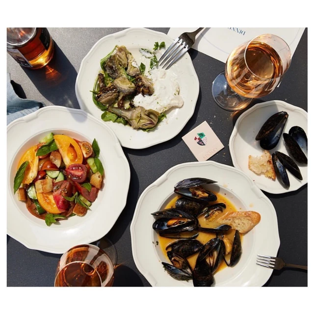 Overhead view of four plates featuring artichoke salad, tomato salad, mussels with broth and bread, and mussels in shells next to wine glasses.