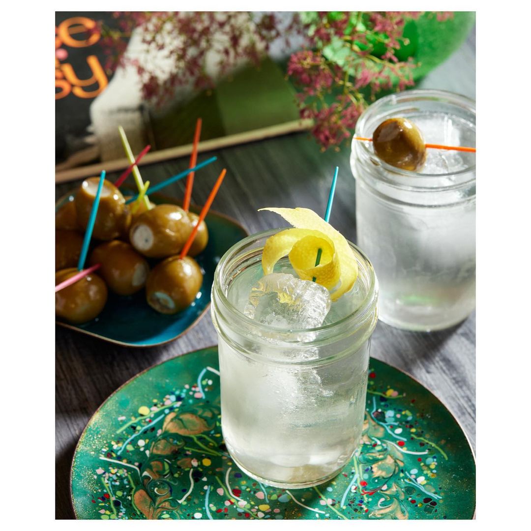 Two glasses of gin with lemon peel and olives on ice, served on decorative green plates with colorful accents and floral background.