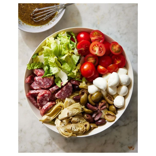 Overhead shot of an Italian salad with salami, mozzarella, artichoke hearts, olives, lettuce and tomatoes in a white bowl, dressing aside.