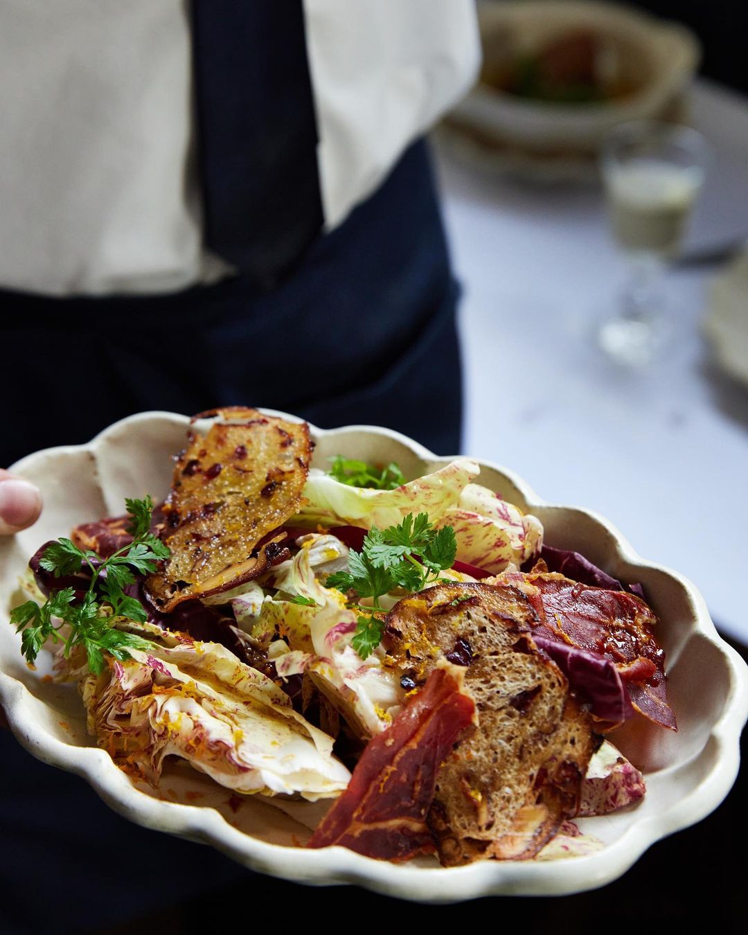 Salad served on decorative plate, featuring mixed greens, toast, and cured meat, held by a person in a restaurant setting.