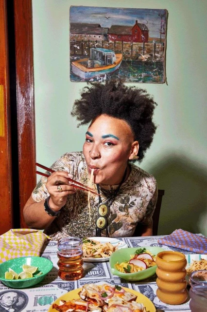 Adult female with Afro hairstyle eating noodles with chopsticks at a table covered with plates of food and a dollar-patterned tablecloth indoors.