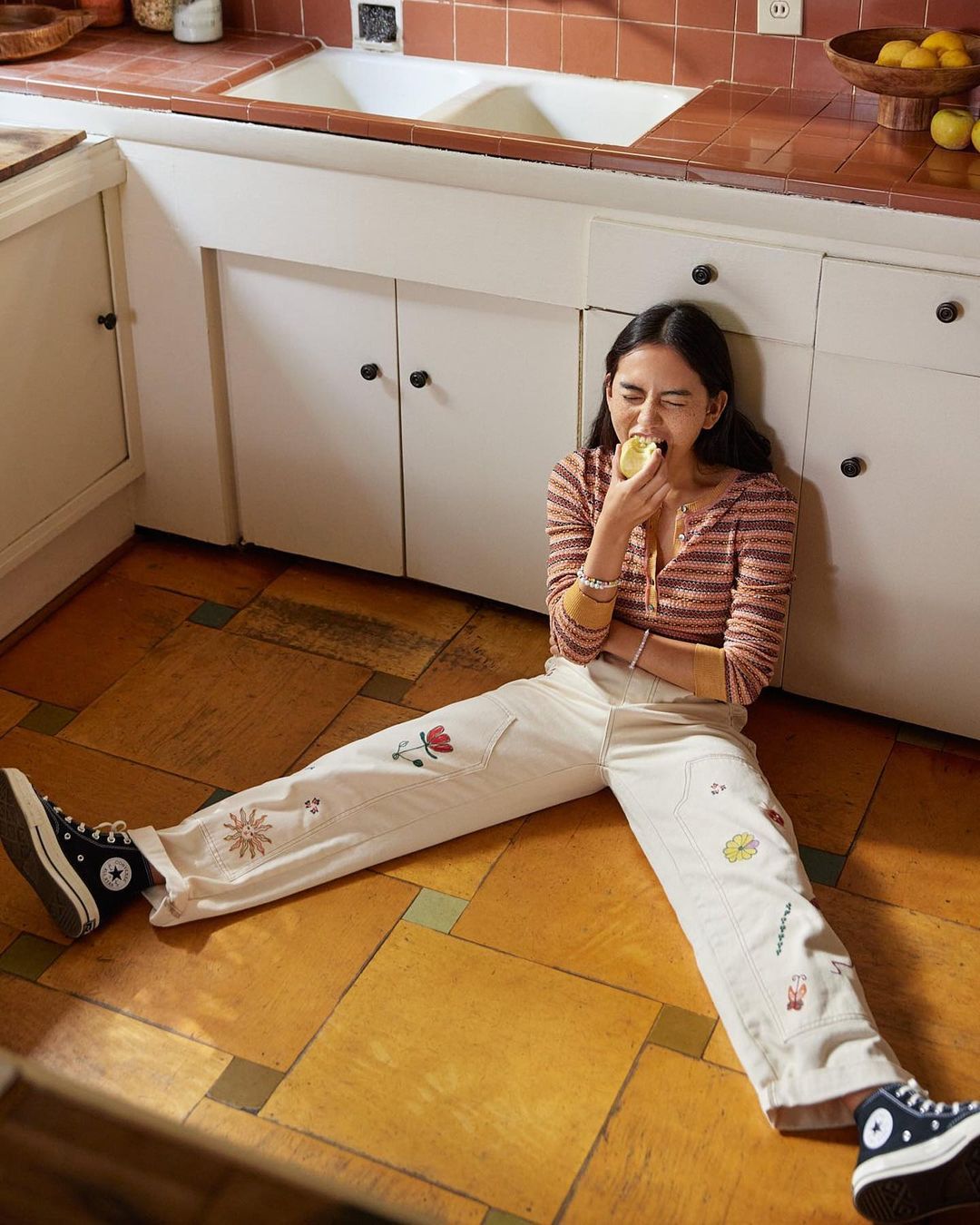 Young woman in striped shirt and patterned pants sits on a tiled kitchen floor, eating fruit near white cabinets and a terracotta countertop.