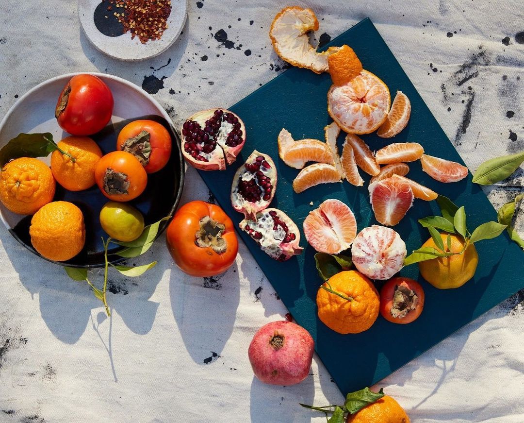 Still life of oranges, tangerines, persimmons, and pomegranates arranged on a table with a white tablecloth and a blue cutting board.