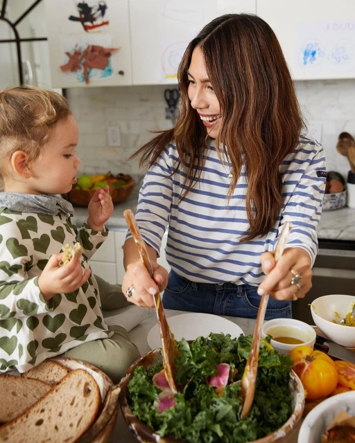 Woman and child preparing a salad with wooden spoons in a bright kitchen. Sliced bread and various ingredients surround the bowl.