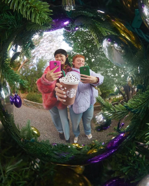 Two smiling women take a selfie with a Starbucks drink in a Christmas ornament reflection, surrounded by green pine needles.