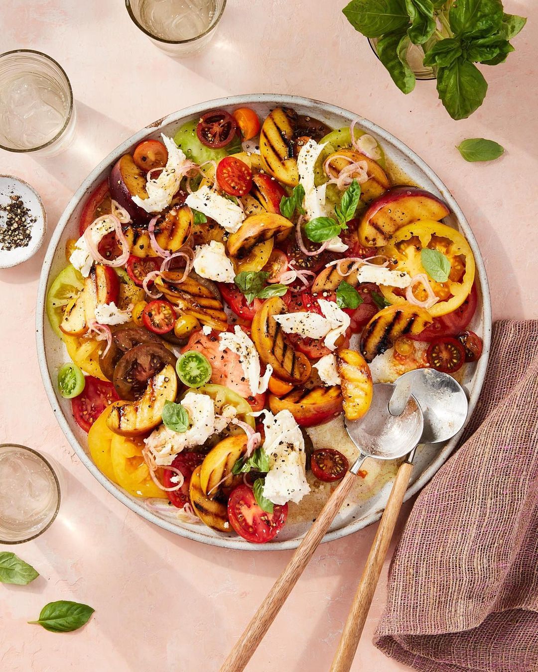 Overhead shot of a vibrant summer salad with heirloom tomatoes, grilled peaches, mozzarella, and basil on a large platter, fresh and colorful.