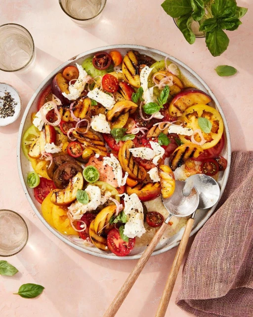 Overhead shot of a vibrant summer salad with heirloom tomatoes, grilled peaches, mozzarella, and basil on a large platter, fresh and colorful.