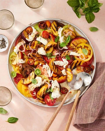 Overhead shot of a vibrant summer salad with heirloom tomatoes, grilled peaches, mozzarella, and basil on a large platter, fresh and colorful.