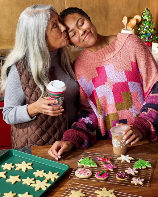 Two women, one kissing the other's cheek, stand at a counter with cookies, drinks, and a Christmas tree visible indoors.