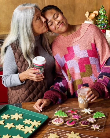 Two women, one kissing the other's cheek, stand at a counter with cookies, drinks, and a Christmas tree visible indoors.