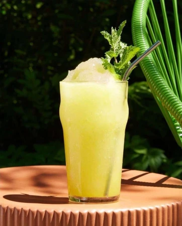 A refreshing lemon slush drink with a metal straw sits on a brown table, with green foliage in the blurry background.
