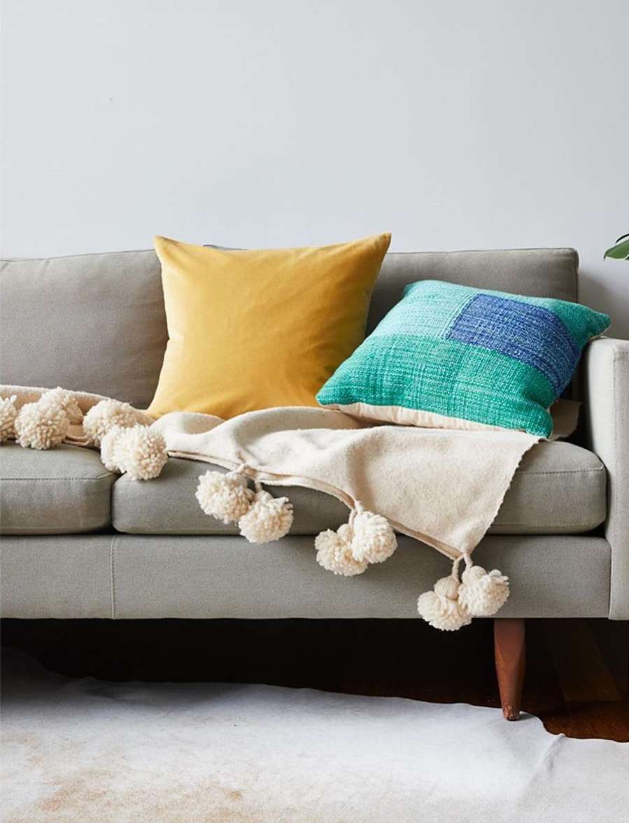 Interior photograph of a grey sofa with two pillows, one yellow and one blue and green, and a pompom blanket, set in a home.