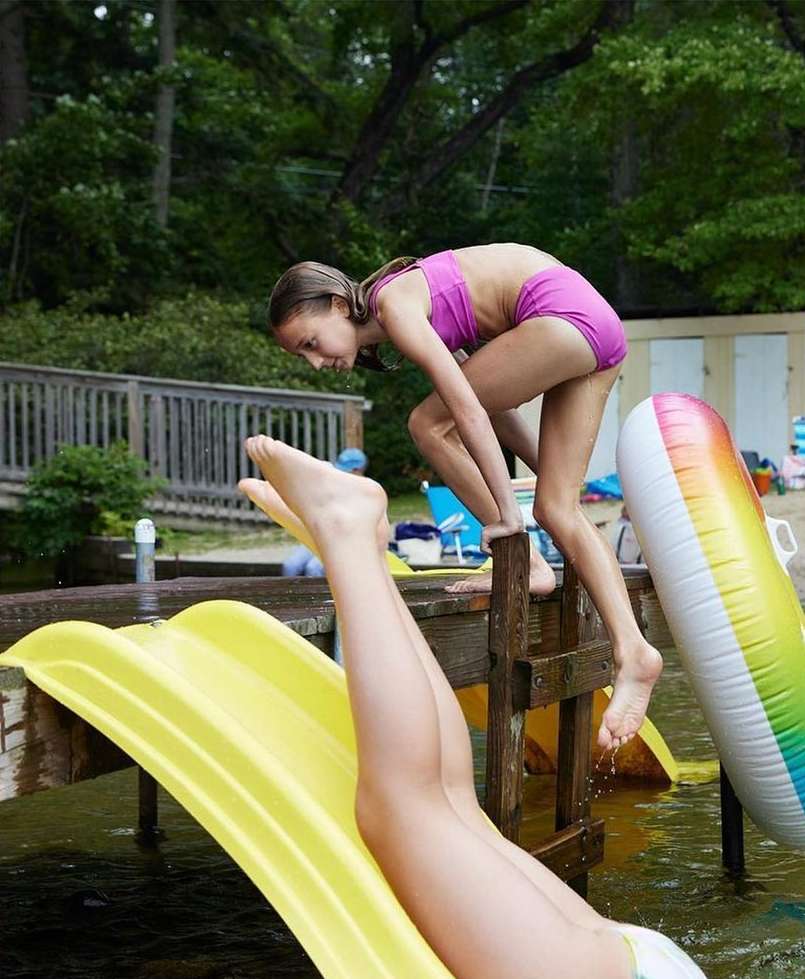Young girl in a purple swimsuit climbing a wooden dock ladder with water slide and inflatable tube by the lake on a summer day.