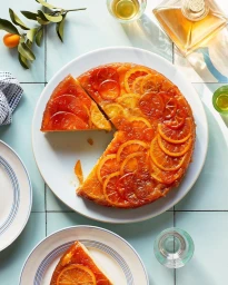 Top-down view of a glazed citrus upside-down cake decorated with sliced oranges on a white plate set on blue tile.
