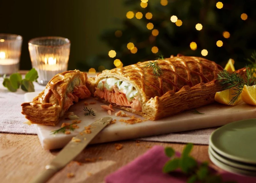 Salmon Wellington pastry with dill and lemon wedges on a marble board, set on a wooden table, Christmas lights in the background.