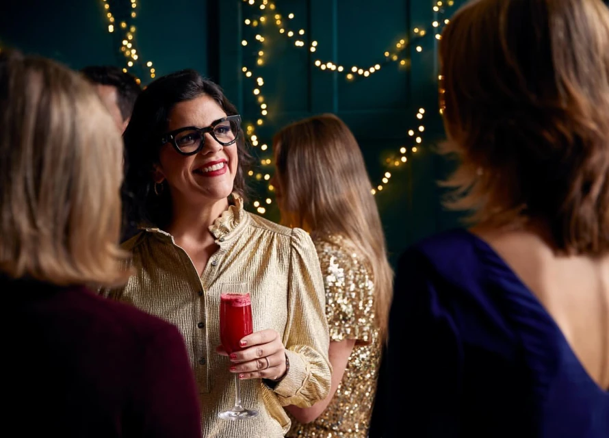 Woman smiles at party holding drink in front of dark green wall with string lights and other partygoers in the background.