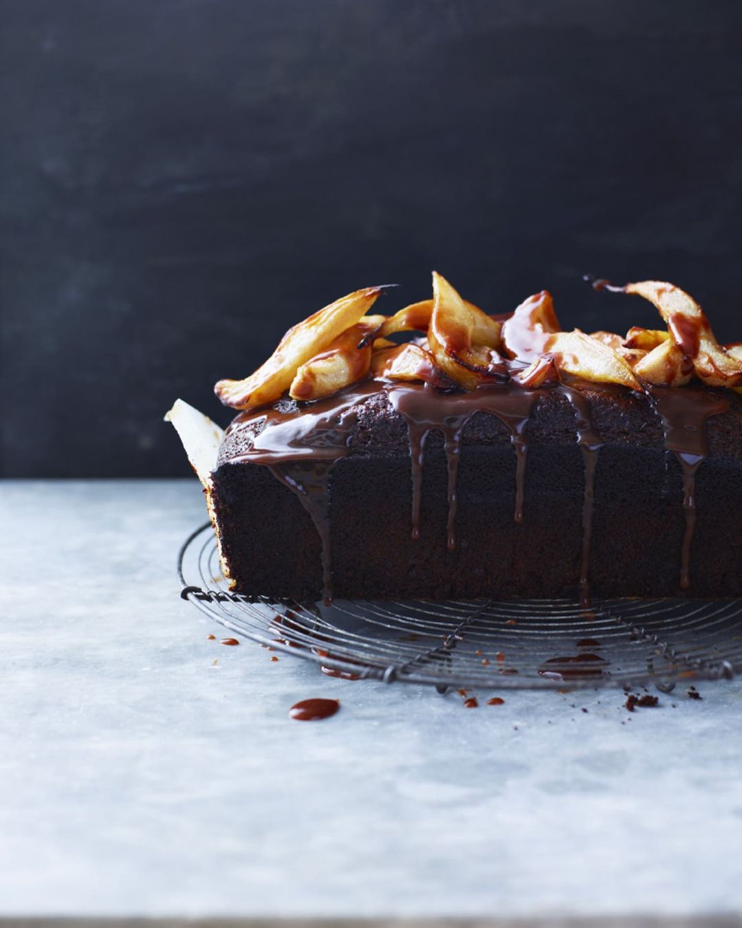 Close-up of chocolate loaf cake topped with pears and chocolate sauce, sitting on a wire rack against a dark background.